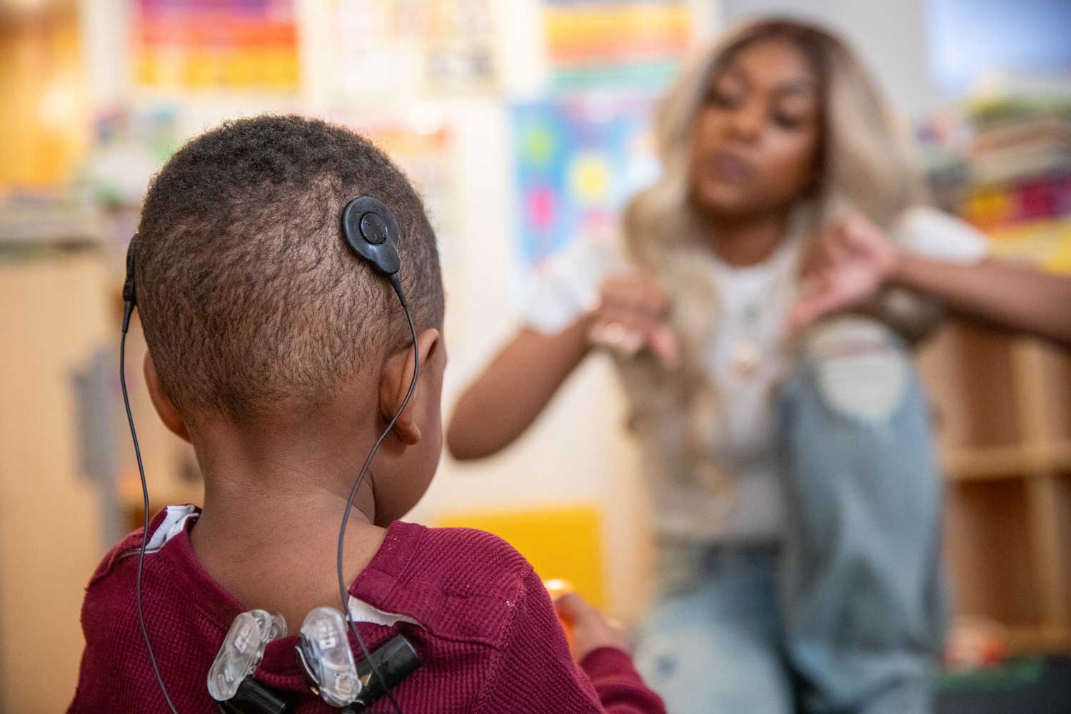 Amir, a toddler, wears two Cochlear implants on the back of his head. In the background, his mother Alisa communicates with him through sign language. He has a cochlear implant, as he became deaf after an infection. They are both of African American descent.