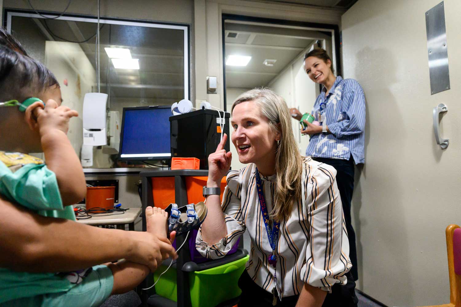 Audiologist Sarah Coulthurst and speech pathologist Annie Sager smile as they test the hearing of a baby patient who sits on his mother's lap and wears a set of headphones.